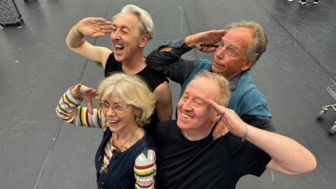 Three grinning men and one grinning woman stand and give a salute in a two-by-two formation. They are all wearing casual clothing and appear to be on a stage during a rehearsal.
