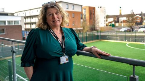 A woman with long blonde hair, glasses on top f her head, a green dress and a lanyard, smiles as she leans on a railing. She is standing on a balcony with a 3G football pitch behind her.