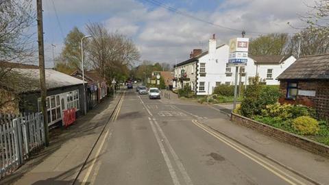 Station Road in Maghull on a dry day with Maghull Railway Station on the right and some cars travelling up the road.