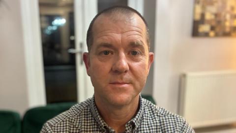 A head-and-shoulders shot of a man wearing a check shirt, looking expressionless directly at the camera.