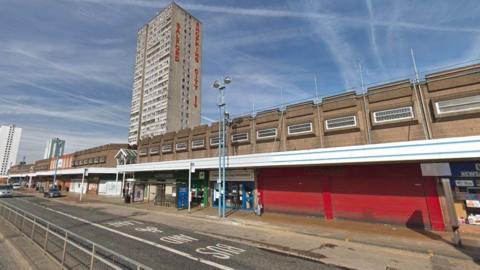 A tower block can be seen in the distance with the words Salford Shopping City on the side in red lettering. There is a bus stop in front of the shopping centre and rows of shops on the ground level. 