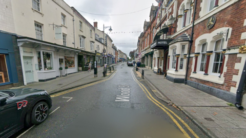 A GoogleMaps image of a street with shops either side and a few parked cars. White lettering reads 'Wood Street' on the road.