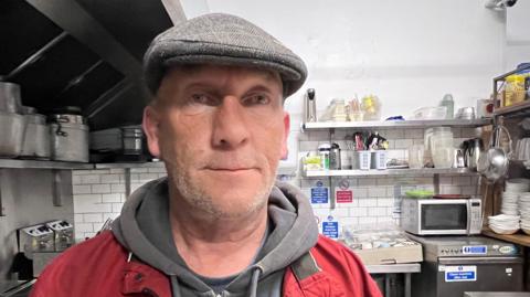 Andrew Talbot looking at the camera while being interviewed at We Are Humans headquarters. He is wearing a flat cap and layers of warm clothes. Food and hygiene supplies could be seen behind him.