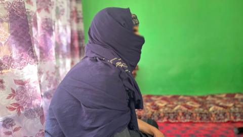 A man is seen wrapped in dark cloths, sitting on an Afghan rug with a green wall in the background