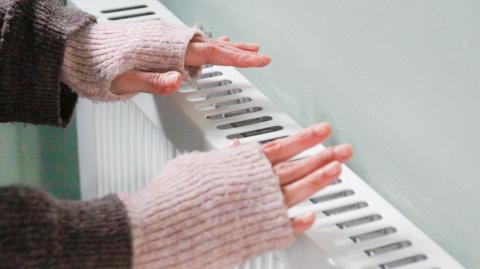 Two hands with with pink fingerless gloves being warmed over a wall radiator