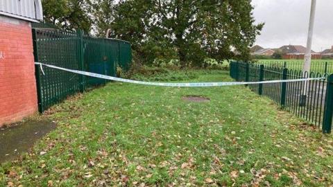 Police tape cordons off an area of grass spotted with leaves, with the tape attached to two green metal gates. 