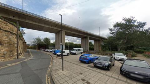 The small car park on Pottery Lane. A number of vehicles are parked in the car park, which sits underneath a large concrete bridge. Pottery Lane is on the left-hand side of the picture and runs alongside a high brick wall.