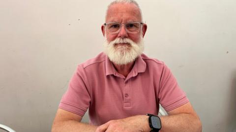 A man with a white beard and glasses sits at a table, wearing a pink polo shirt, with his arms resting in front of him.
