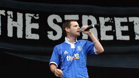 Mike Skinner in 2004 on stage wearing a blue Flybe football top, holding a microphone. Behind him is a large banner with bold, distressed white text reads “THE STREETS”.