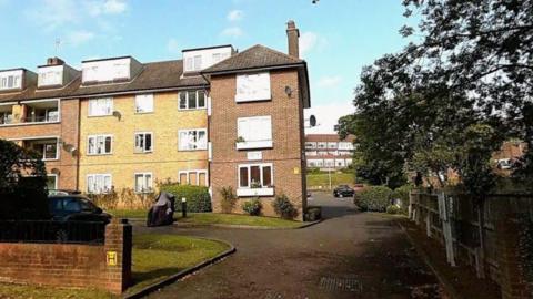 Google street map view of Kingfisher Court, off Queensmere Road, at three-storey brick apartment building with dormer windows and a paved driveway, bordered by grass, shrubs, parked vehicles and mature trees under a blue sky.