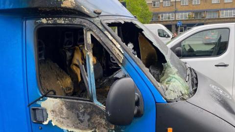 The front of a blue van is charred black and damaged with missing paint and smashed glass.
