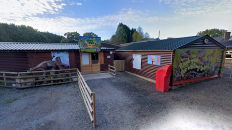The entrance of a theme park, Hoo Zoo, with colourful signage on a series of single-storey wooden buildings. There is a fence to guide queues and a sign saying "entrance" above a door.