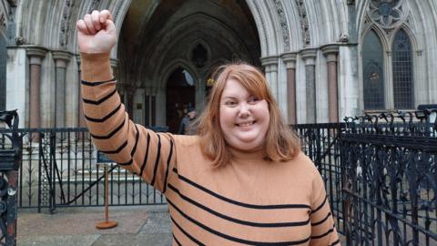 Former Liberal Democrat parliamentary candidate Natalie Bird stands outside court. She is wearing a brown and black striped jumper and is holding her fist in the air, smiling. She has mid-length ginger hair.