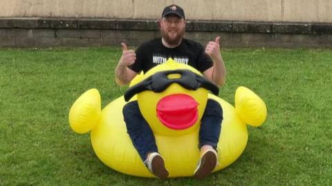 Liam Birkett sits astride his inflatable duck on a patch of grass with his thumbs up. He is wearing dark jeans, a black tshirt and baseball cap and has a brown beard