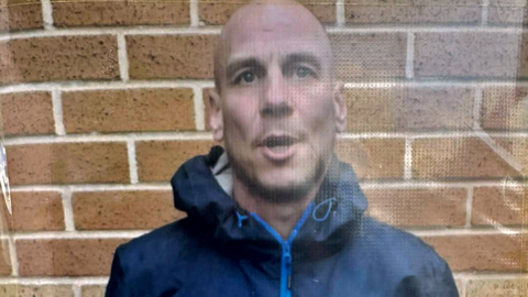 A bald man, who is wearing a dark blue coat with the hood down, has his mouth partially open as he stands in front of a wall.