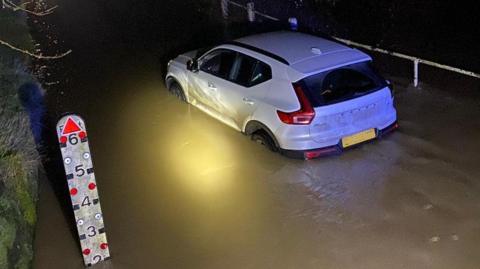 A white car is stuck in flood water. The water is half-way up the tyres and there is a measuring gauge at the side saying that the water is 2ft deep (0.6m)