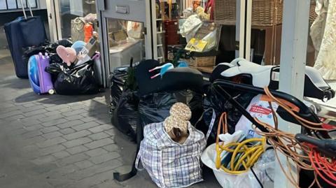 A shop front at night-time with grey concrete flooring outside. Against the windows of the shop, piles of fly-tipping can be seen including suitcases, a lawnmower, teddies, car seats, wires and many black bin bags. 