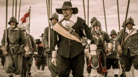 An English Civil Ware enactment, showing an image of several soldiers, wearing historic outfits, armour, holding sticks. They all have hats or helmets on. 