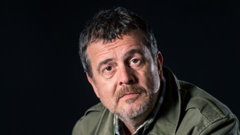 A close-up of a man with dark grey hair looking into the camera. He wears a dark-green jacket and is in front of a dark background.