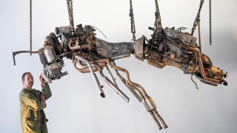 Joe Rush, wearing a brown overcoat, stands next to one of his works, a giant bee made using three old engines. It hangs in the air from several chains in front of a plain white wall.