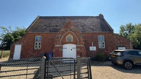 A old brick building with white doors in the middle of a gravel driveway. A black metal gate and grey car are nearby.