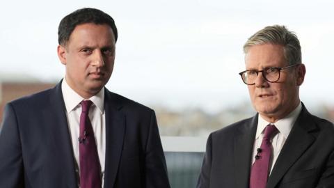 Two  men stand side-by-side posing for a photograph. They are both wearing dark suits, white shirts with ties. 