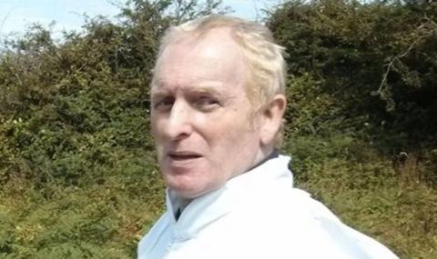 A man with light blonde hair, wearing a white shirt, looks at the camera. There are thick bushes in the background.