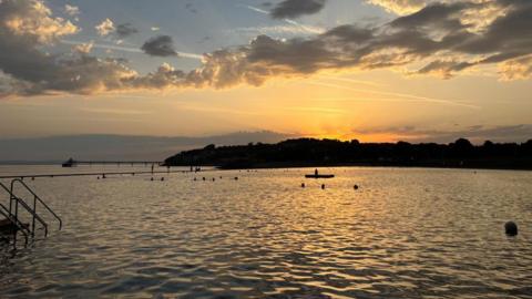 The sun is seen rising over Clevedon Marine Lake at dawn. The water is calm and the sun is lighting up the horizon beyond the pier with bright orange hues. There are some swimmers in the water, visible only as silhouettes.