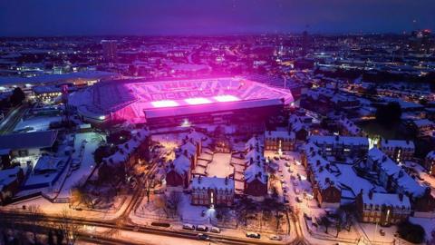 An aerial view of Birmingham City Football Club ground, St Andrews. It is surrounded by snow-covered buildings in the city centre and the pitch can be seen lit up in a bright pink colour. 