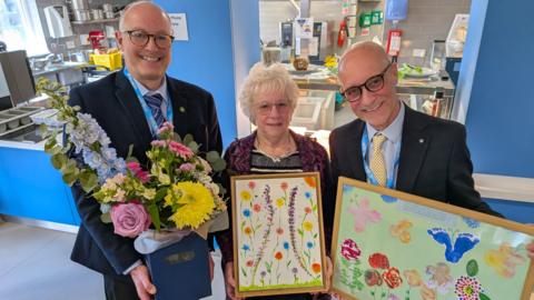 Three people are smiling at the camera. A man on the left is bald with glasses and is wearing a black suit. He is holding a large box of flowers that are pink, yellow and blue. A woman in the middle has short white hair, glasses and is wearing a black top and red cardigan. She is holding a framed painting of colourful flowers. On the right is another man who is bald with glasses, who is wearing a light blue shirt and black suit jacket. He is holding a painting with flowers and children's footprints on it 