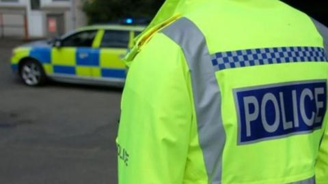 Stock image of a police officer pictured from behind, wearing a yellow high-viz police jacket. They are standing close to a police car.