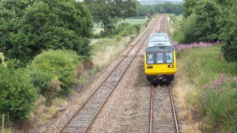 A small GWR train on a track. There is another train track on the left. Around the lines are green grass and trees with green leaves. 