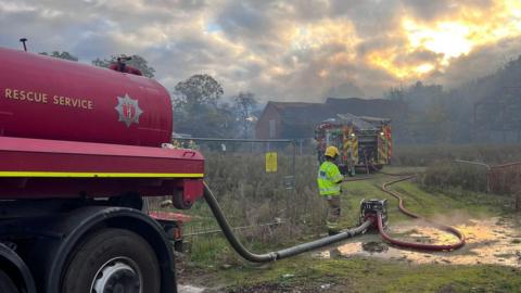 A red water carrier with Warwick Fire and Rescue Service's emblem can be seen in the foreground, with a hose attached to a pump and a fire engine in the distance. there is a firefighter stood next to the pump, wearing a high visibility jacket and a yellow helmet.