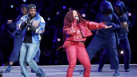 Kendrick Lamar and SZA performing at the Super Bowl. Kendrick Lamar is wearing a black cap and t-shirt, a jacket and blue jeans. SZA is wearing red trousers and a red jacket and has long red hair. 
