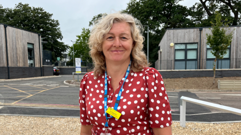 Jo Segasby is outdoors. She is looking directly at the camera and smiling. She is wearing a red and white polka dot white blouse and a blue NHS lanyard round her neck.