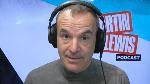 Martin Lewis wearing a grey jumper and headphones, sat in a studio. 