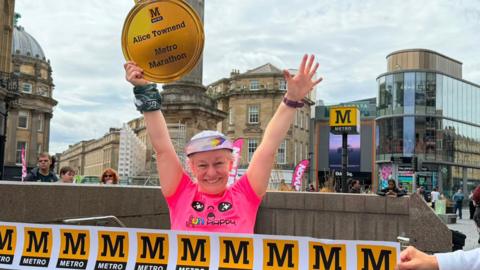 Alice Townsend smiling into the camera with her arms up in the air. She is wearing a colourful cap with the brim standing up and has white hair peeking through. She is wearing a bright pink t-shirt and is holding a large cardboard medal in one of her hands which reads "Alice Townsend Metro Marathon". People are holding a white finishing line banner in front of her with the yellow Metro logo, which is a black capital M in a yellow box. The bottom of the stone Grey's Monument is behind her, as well as a large Metro sign. The stone buildings of Grainger Street are also behind her along with the glass-fronted Eldon Square. 