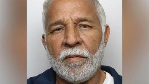 Piran Ditta Khan frowning with a light grey beard and light grey hair. He is stood in front of a light grey background and wearing what appears to be a white t-shirt and blue jacket.