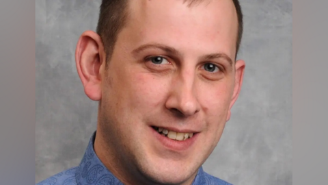Peter Little wears a blue paisley shirt and stands for a portrait photo in front of a grey screen. He has brown hair which is receding and blue eyes. 