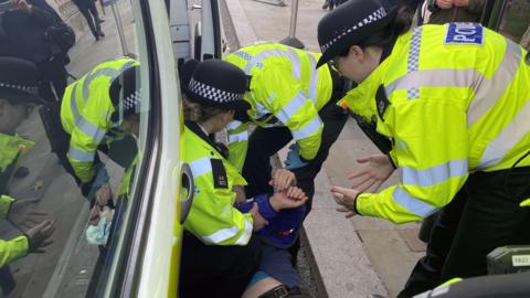 Three police officers in uniform arrest a figure lying down on the ground between a kerb and a vehicle. The individuals' face is hidden by their outstretched arms and the uniform of one of the officers.