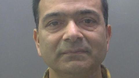 A custody image of Pramod Dhakal. He has short dark hair and is looking into the camera. He has a yellow and black checked shirt collar.