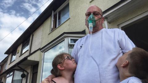 Rob Hill wearing an oxygen mask, embracing two of his children