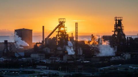 The steelworks in Port Talbot at sunset. White steam rises from the chimneys. 