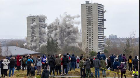 blocks of flats are destroyed by explosions as people in the foreground watch