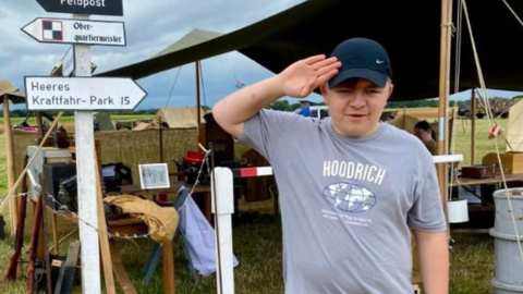 A young man with a navy blue cap and a grey tshirts saluting in front of a tent and a sign post