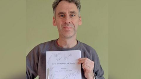 Robert Wilfort, who plays Jason in Gavin and Stacey, smiles at the camera holding the Christmas Day episode finale script which has been signed bby all the main cast. Robert has grey hair and is wearing a grey jumper. 