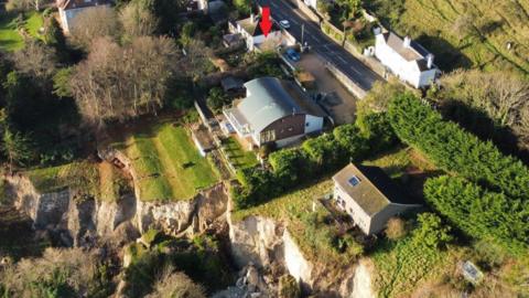 A drone shot of the houses near the edge of the landslip. The house in question is a modest semi-detached further back from the cliff than the others, nearer to the road.