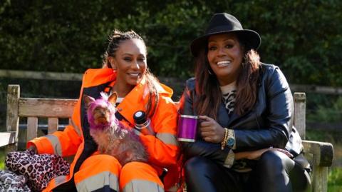 Mel B hold a small dog and sits with Alison Hammond on a park bench