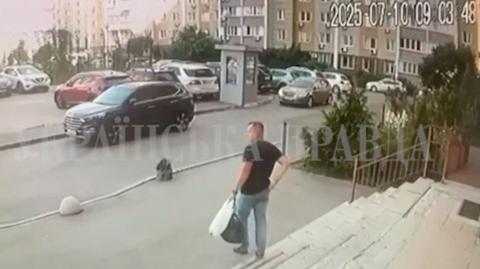 A man in jeans and a dark t-shirt holding a plastic bag and a holdall in one hand walks out of a building towards a car park in Kyiv.