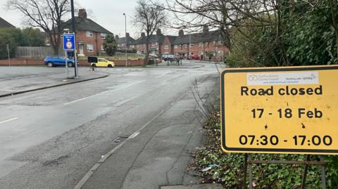 A yellow road sign with details of a road closure, is positioned at the side of a road with a junction visible in the distance.